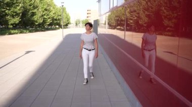 Şık bir kadın, güneşli bir şehir kaldırımında güvenli adımlarla yürüyor şık kıyafetlerini, hareketli şehir hayatının ve modern mimarinin canlı arkaplanına karşı zekice sergiliyor.