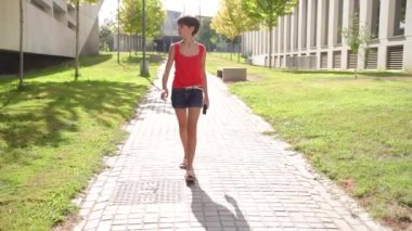 Kırmızı atletli ve şık şortlu kendine güvenen genç bir kadın yeşil ve modern mimarisi zengin güzel bir parkta güneşli bir yolda yürüyor ve manzarasını güzelleştiriyor.