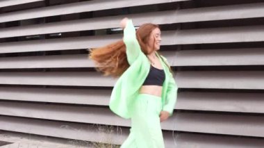 Canlı genç bir kadın, enerji ve neşe saçıyor, çarpıcı yeşil bir takım elbise içinde çağdaş bir mimari binanın önünde neşeyle dans ediyor, dinamik ve kaygısız ruhunu sergiliyor.