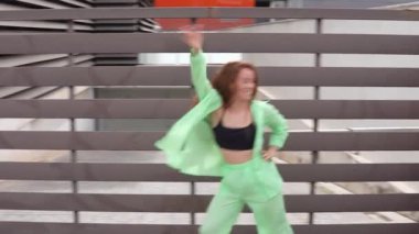 Canlı bir şehir sahnesinde, parlak yeşil eşofmanlı bir kadın modern bir mimari çitin yanında enerjik bir şekilde dans ediyor. Dinamik hareket ve renkli kıyafetler enerjik bir atmosfer yaratır.