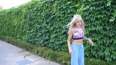 Genç, sarışın bir kadın canlı bir sarmaşık duvarın önünde enerjik bir şekilde dans ediyor, doğayı kucaklıyor, açık hava aktivitelerinden ve yaz havasından keyif alıyor. Onun kaygısız ruhu yeşilliğe güzellik katıyor.