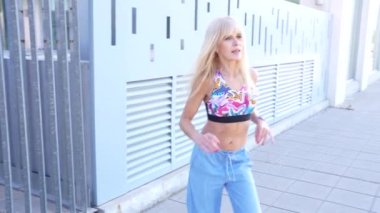 Sarışın bir kadın modern bir binanın önünden şık bir kıyafetle geçerek güneşli günün tadını çıkarıyor. Moda kıyafetleri içinde şık ve rahat bir hava yayıyor, özgürlük ve yaz havasını somutlaştırıyor.