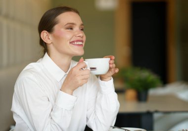 Gülümseyen iş kadını kafeteryada nefis aromatik bir kahvenin tadını çıkarıyor. Elinde fincanla tek başına oturuyor.