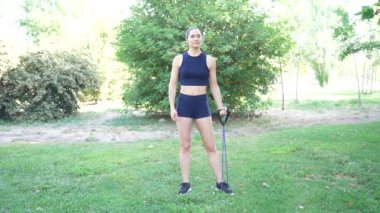 Parkta, bir kadın çeşitli egzersizler için direnç bandı kullanır. Jimnastik, kardiyo ve kas tonlamasını geliştirmek için. Kapsamlı bir açık hava spor programı yaratır.