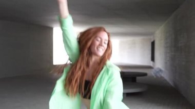 Uzun kızıl saçlı genç bir kadın modern bir şehir tünelinde enerjik bir şekilde dans ediyor, neşe ve özgürlük saçıyor. Yeşil ceket minimalist beton çevreye biraz renk katıyor..