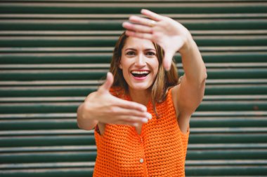 Neşeli Latin kadın kameraya bakıyor ve gülümsüyor şehir sokaklarında parmaklarıyla çerçeveyi şekillendiriyor.