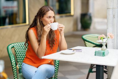 Asfalt kafede rahatça oturup kahve içen Latin bir kadının üç çeyrek boy fotoğrafı.