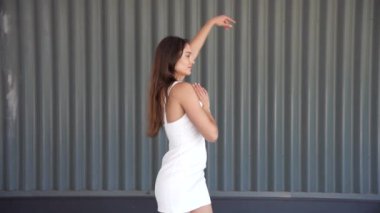 Şaşırtıcı derecede şık, genç bir kadın şık beyaz bir elbise içinde kendine güvenli bir poz veriyor, modern metalik bir arka plana zarif bir şekilde yerleştirilmiş, stil ve zarafet duygusunu güzel bir şekilde sergiliyor.