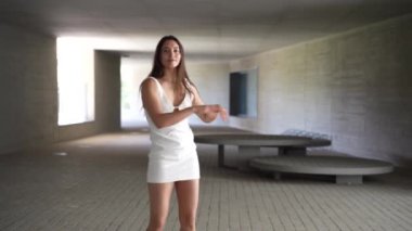 Şık beyaz bir elbise giyen genç bir kadın modern, aydınlık bir ortamda zarifçe poz veriyor, izleyicilerin ilgisini ve hayal gücünü gerçekten cezbeden büyüleyici bir atmosfer yaratıyor.