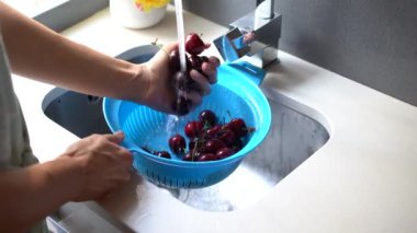 Yüksek açılı, yüzü olmayan bir kadının, mutfak lavabosundaki musluktan akan suyun altında kirazları plastikle yıkarkenki statik görüntüsü.
