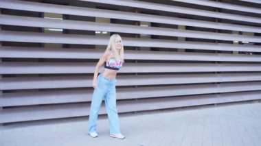 Hareketli bir şehir ortamında, dinamik ve şık bir sarışın kadın, atletik kıyafetler giyer, kendinden emin bir şekilde canlı dans hareketleri sergiler. Yetenek ve dışavurumculuk, enerji ve pozitiflik yayar.