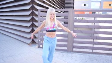 Şehir ortamında, canlı sarışın bir kadın enerji dolu bir bluz ve kotla dans ediyor, spontanelik ve neşe saçıyor. Sahne neşeli ve neşeli, tasasız ve genç bir atmosferi somutlaştırıyor.