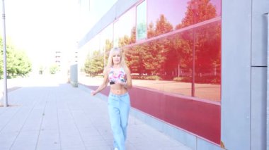 Canlı bir şehir manzarasında, genç sarışın bir kadın modern gökdelenler arasında geziyor, enerjik bir şehir havası yayıyor. Gündelik tarzı, canlı şehrin çağdaş atmosferiyle uyuşuyor.