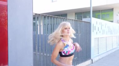 Şaşaalı, uzun sarı saçlı şık bir kadın modern bir binanın önünden geçerek göz alıcı kırmızı panelleri, renkli bluzu ve kotu ile kentin moda yaşam tarzını yansıtıyor.