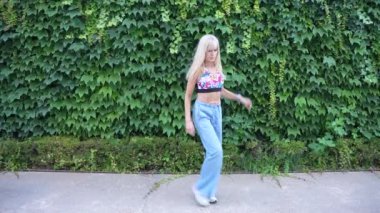 Uzun sarı saçlı, şık bir genç kadın, çiçekli bir bluz ve wideleg kottan oluşan modaya uygun giyinmiş, fildişi kaplı bir duvara karşı güvenle poz veriyor. Modern moda ve kendine güven yayıyor.