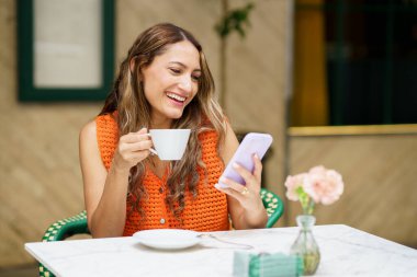Kafeteryada cep telefonu kullanırken gülümseyen mutlu ve rahat bir Latin kadının ön fotoğrafı.