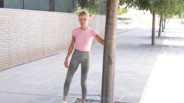 Atletik giyinen bir kadın, dışarıda, ağacın yanında bir direniş bandı kullanır, doğal ortamında zindelik gösterir ve egzersiz ve fiziksel refah yoluyla sağlıklı bir yaşam tarzını teşvik eder.