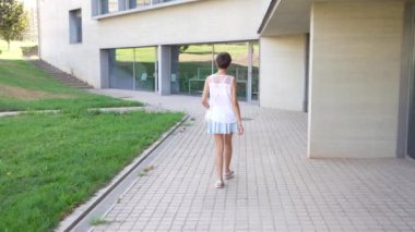 Genç bir kız, çağdaş mimari ve yemyeşil çimlerle çevrili, kaygısız çocukluk anlarında macerayı ve keşfi somutlaştıran asfalt bir yolda neşeyle yürüyor.