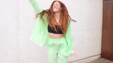 A vibrant young woman joyfully dances outdoors against a white and brown wall in a bold green outfit with black sports bra, her hair flowing as she moves with energy and enthusiasm