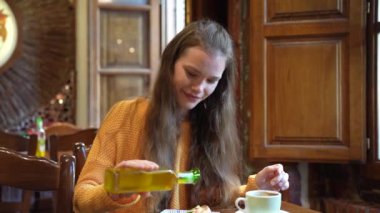 Uzun saçlı genç bir kadın ahşap bir masada oturuyor, sıcak bir kafede kahve bardağını tutarken gülümsüyor.