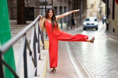 Göz kamaştırıcı kırmızı elbiseli şık ve şık bir kadın hayat ve renklerle dolu büyüleyici ve pitoresk bir şehir caddesinin ortasındaki korkulukta neşeli bir poz verir.