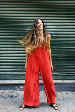 Şık kırmızı bir tulum giymiş neşeli ve hayat dolu bir kadın, zarif ve kendine güvenen yeşil bir zemine karşı, modaya ve kendini ifade etmeye olan sevgisini güzel bir şekilde sergiliyor.