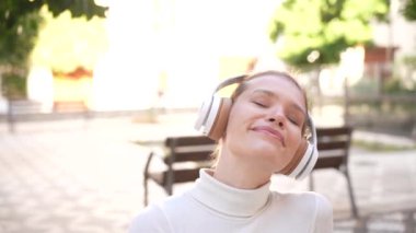 Huzurlu bir parkta, zarif kulaklıklı neşeli genç bir kadın müziğine dalmış, açık havanın tadını çıkarırken doğanın güzelliğini yansıtan ışıl ışıl gülüşü