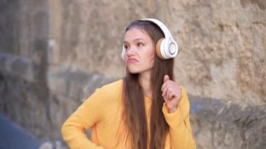 Güneşli bir şehir geçidinde, uzun saçlı genç bir kadın, rahat, kaygısız bir keyif ve özgürlük hissini yansıtan rahat sarı bir kazak giyip moda kulaklıklarıyla müzik dinlemekten hoşlanıyor.