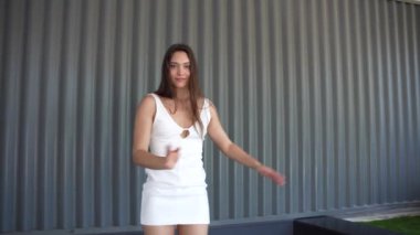 Zarif genç bir kadın, akıcı beyaz bir elbiseyle sanatsal havayı geliştiren, enerji dolu dinamik bir atmosfer yaratan dokulu bir duvar tarafından çerçevelenmiş etkileyici dans figürleri sergiliyor.