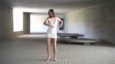 Kendine güvenen ve özgüvenli bir kadın, çarpıcı beyaz bir elbise giymiş, zarif bir şekilde modern ve şık bir mekanda süzülüyor, doğal güzelliği ve cazibesiyle etrafındaki herkesi büyülüyor.