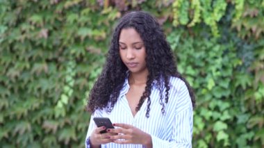 Kıvırcık saçlı genç bir kadın dijital dünyaya dalmış, çarpıcı bir yeşillikle çevrili, modern teknolojiyi doğal sakinlikle harmanlayan akıllı telefonuna odaklanmış durumda.