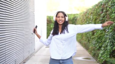 Neşeli ve kaygısız genç bir kadın akıllı telefonuyla çaldığı müziğe dalıp şık kulaklıklar takarak açık havada canlı ve canlı bir atmosferi ön plana çıkarıyor.