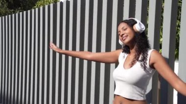 Moda kıyafetleri içinde genç bir kadın, modern bir çitin yanında duruyor, en sevdiği müziğin tadını çıkarıyor ve neşeli enerjisiyle eşleşen açık havada mutluluk saçıyor.