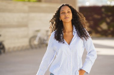 Güzel kıvırcık saçlı şık bir genç kadın, kendine güvenen bir şekilde dışarıda dolaşır. Modaya uygun mavi ve beyaz çizgili bir gömlek giyer. Etrafındakileri büyüleyen bir zarafet ve duruş duygusu somutlaştırır.