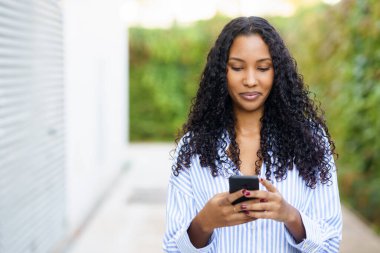 Sakin bir açık havada, kıvırcık saçlı genç bir kadın akıllı telefonuyla gelişigüzel etkileşime giriyor, dijital deneyimiyle meşgul olurken çevresindeki güzelliğin tadını çıkarıyor.