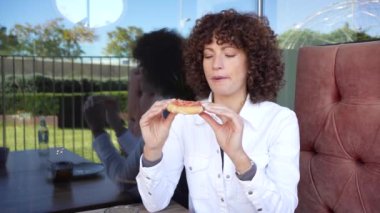 Kıvırcık saçlı genç bir kadın, bir kafede oturur, düşünceli bir şekilde elinde bir dilim pizzayla, sakin bir atmosferle yemeğini yansıtır.