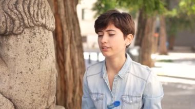 Kısa saçlı genç bir birey düşünceli bir şekilde taş bir heykelin yanında oturuyor, derin bir tefekküre daldırılmış, ve canlı kentsel çevrede yemyeşil ile çevrili.