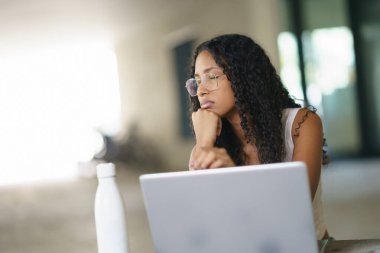 Parlak, çağdaş bir çalışma alanında, kıvırcık saçlı ve şık gözlükleri olan genç bir kadın dikkatle dizüstü bilgisayarına odaklanıyor, bir sonraki stratejik hamlesini ve gelecek planlarını düşünürken görevlerine odaklanıyor.