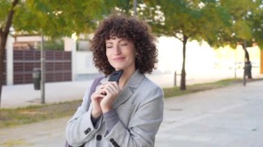 Kıvırcık saçlı neşeli genç bir kadın akıllı bir telefon tutuyor ve gülümsüyor, canlı bir şehir zeminine kurulmuş, yemyeşil ağaçlar ve çağdaş ruhu yansıtan renkli şehir unsurlarıyla.