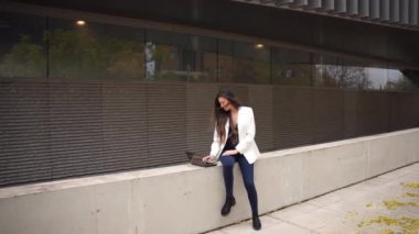 Şık beyaz ceket ve modaya uygun kot giyen şık genç bir kadın çağdaş bir mimari sitede poz veriyor, şık, modern bir arka plana karşı moda anlayışını sergiliyor, kentsel yaşamı somutlaştırıyor.