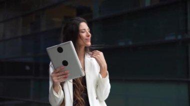 Şık bir kıyafet giymiş bir kadın tabletle meşgul oluyor, canlı bir sanal çağrı boyunca çeşitli duygular sergiliyor açık havada şık, modern bir binanın yanında gün ışığında.