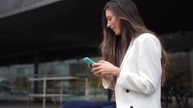 Yoğun bir şehirde, trend beyaz ceketli genç bir kadın dikkat çekiyor, akıllı telefonuyla özümsüyor, günümüz dijital dünyasında modern iletişim ve profesyonelliği somutlaştırıyor.