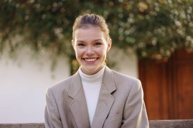 Canlı genç bir kadın kameraya gülümserken özgüven ve sıcaklık saçar, şık bir ceket ve rahat bir balıkçı yaka giyer, güzel bir yeşil zemin ve güneş ışığı altında.