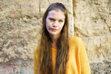 Sarı kazaklı genç bir kadının canlı bir portresi dalgalı saçlarını ve dingin ifadesini dokulu taş bir duvara karşı sergiliyor. Benzersiz cazibesini ve kendine güvenini yansıtıyor.