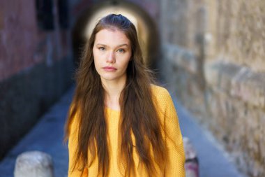 Parlak sarı bir süveter giymiş genç bir kadın, manzaranın genel atmosferini güzelleştiren pitoresk taş duvarlarla dolu tarihi bir ara sokakta güven içinde duruyor.