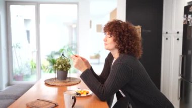 Rahatlamış ve sakin bir kadın mutfak masasında rahat bir şekilde oturur, kahvesinin ve sıcak, doğal ışıkla dolu sıcak ve modern bir mekanda keyifli hafif bir kahvaltının tadını çıkarır.