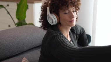 Genç bir kadın kanepesinde dinleniyor, kulaklıklarla müziği dinleyip sıcak bitki çayını yudumluyor, günlük yaşamını zenginleştiren huzurlu ve basit şehir yaşamını kucaklıyor.