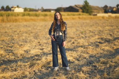 Kendine güvenen genç bir kadın güneşli bir alanda şık kotlar ve canlı bir bluzla duruyor. Neşeli ruhunu ve gençlik enerjisini arttıran resimsel bir manzarayla çevrili.