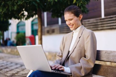 Genç bir kadın laptopunu mutlu bir şekilde açık hava ortamında rahat bir bankta otururken, yemyeşil bir ortam ve neşesini artıran ve işine odaklanmasını sağlayan parlak gün ışığının sıcaklığı ile çevrili bir şekilde kullanıyor.