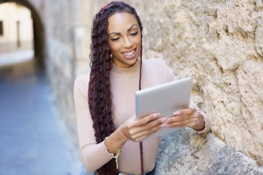Uzun örgülü saçları olan neşeli bir kadın tabletine odaklanmış, modern teknolojiyle tarihi güzelliği harmanlayan, göz alıcı bir sahne yaratan dokulu taş bir duvarın karşısında duruyor.